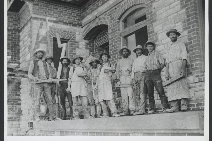 Nine men in work attire standing on a building ledge holding tools, early construction scene.