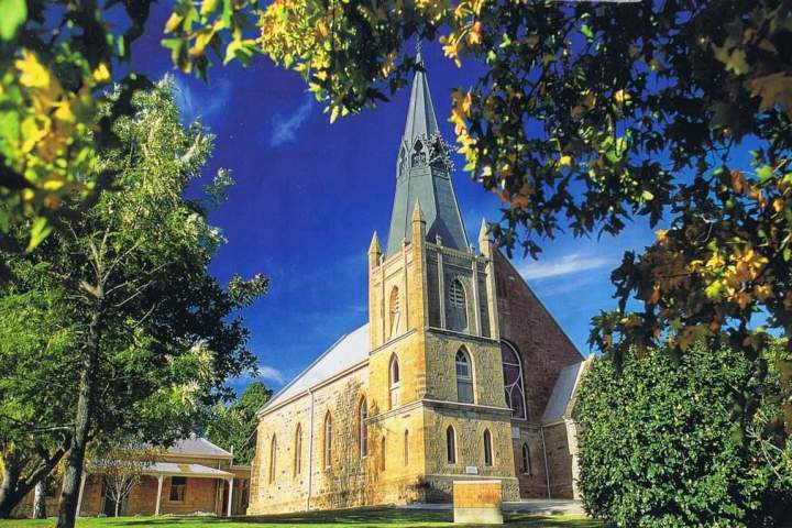 Stone church with a tall spire surrounded by trees under a clear blue sky.