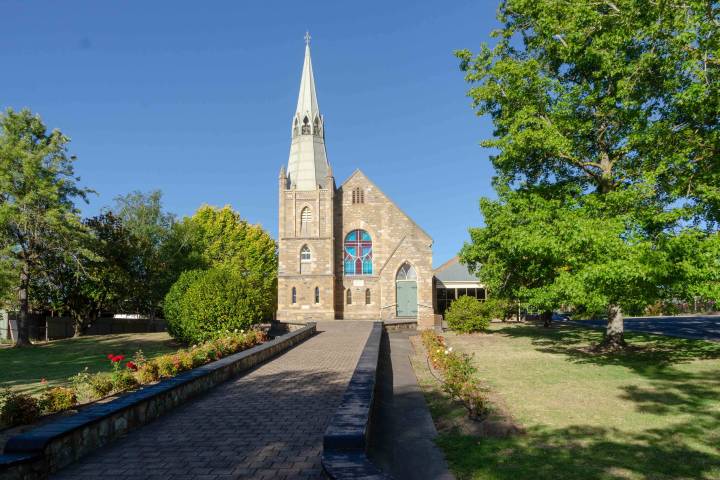 Stone church with tall spire, stained glass window, surrounded by trees and pathway under clear blue sky.