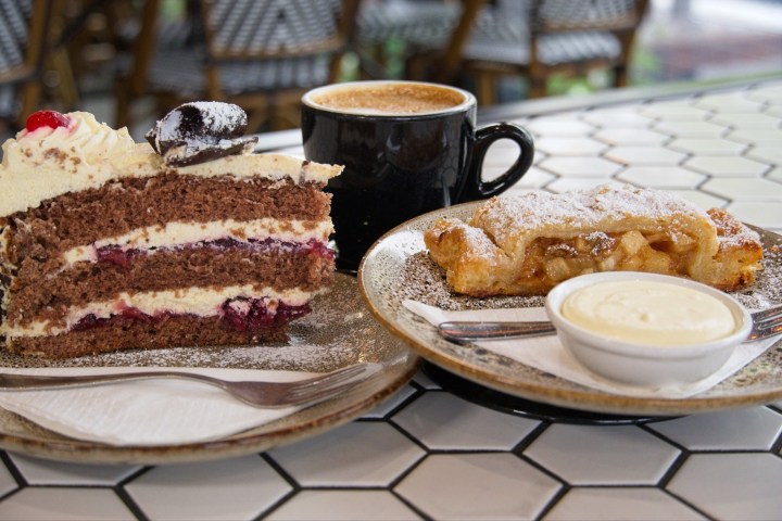 Desserts on a tile table: cake, apple strudel with cream, and coffee in a black mug.
