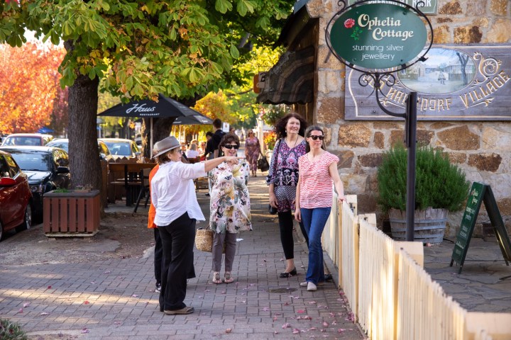 Group of people walking on a tree-lined sidewalk beside a stone building with signs.